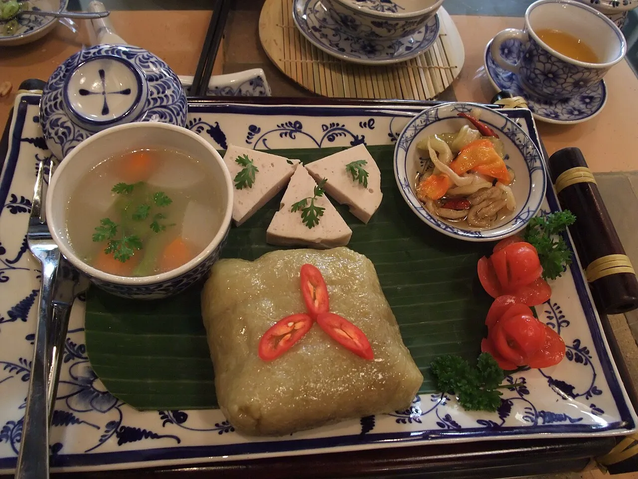 Plateau avec une part carrée de bánh chưng posée sur une feuille de bananier, des tranches de giò lụa, un bol de soupe et une coupelle de légumes marinés