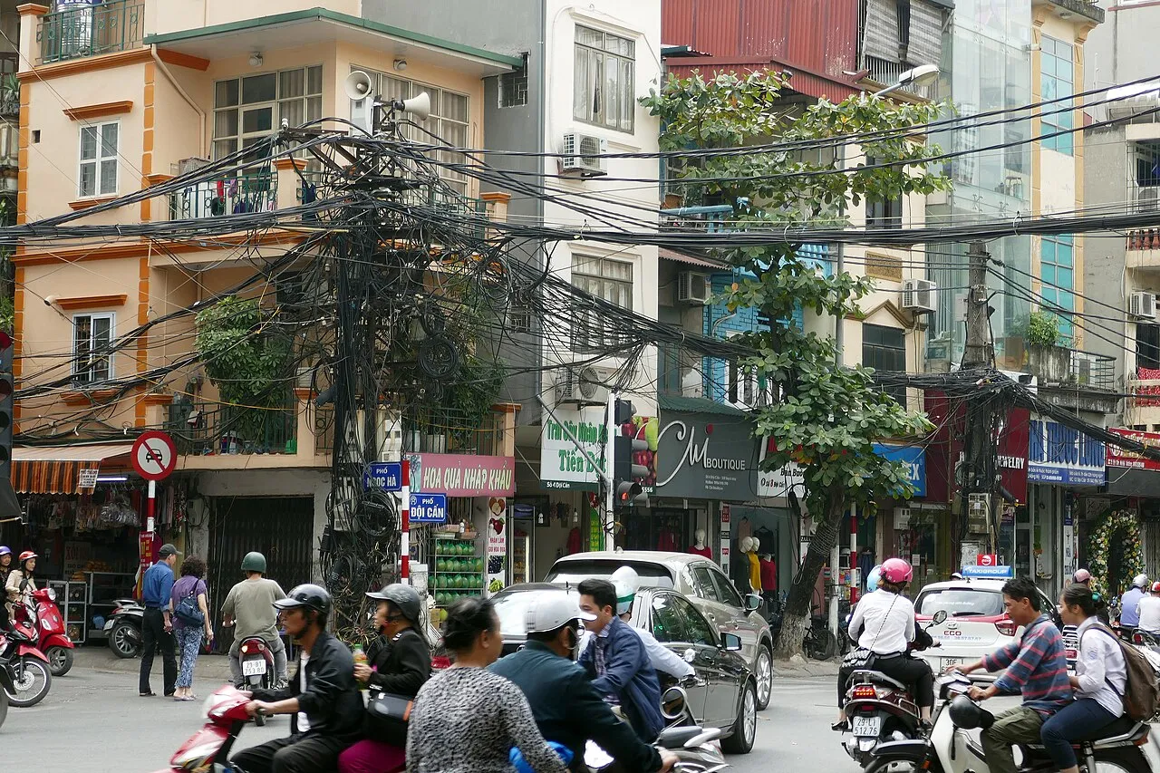 Enchevêtrement de lignes électriques accrochées à un poteau au-dessus d'une rue de Hanoï, avec des motos et des commerces en contrebas