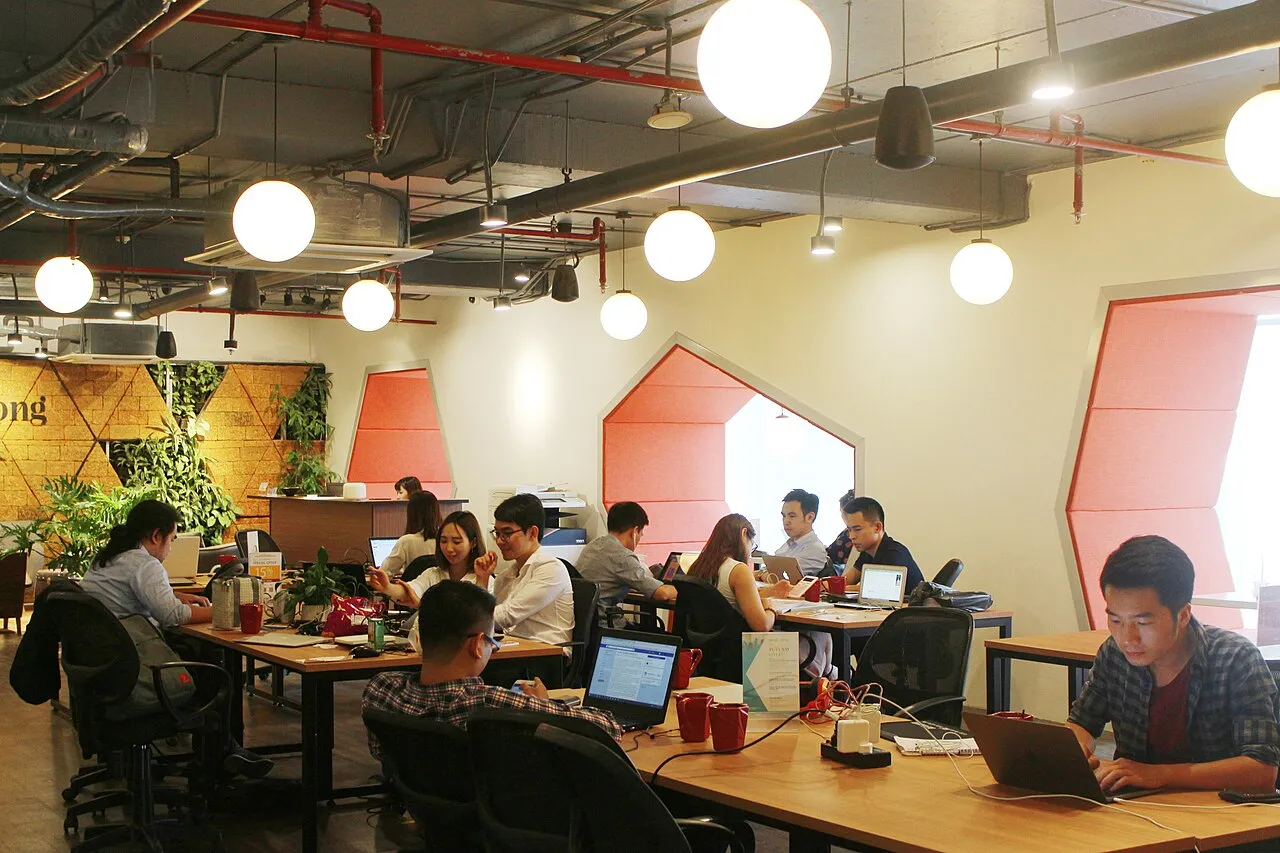 Des personnes travaillent sur leurs ordinateurs portables aux longues tables d'un espace de coworking Toong à Hanoï, sous des lampes rondes