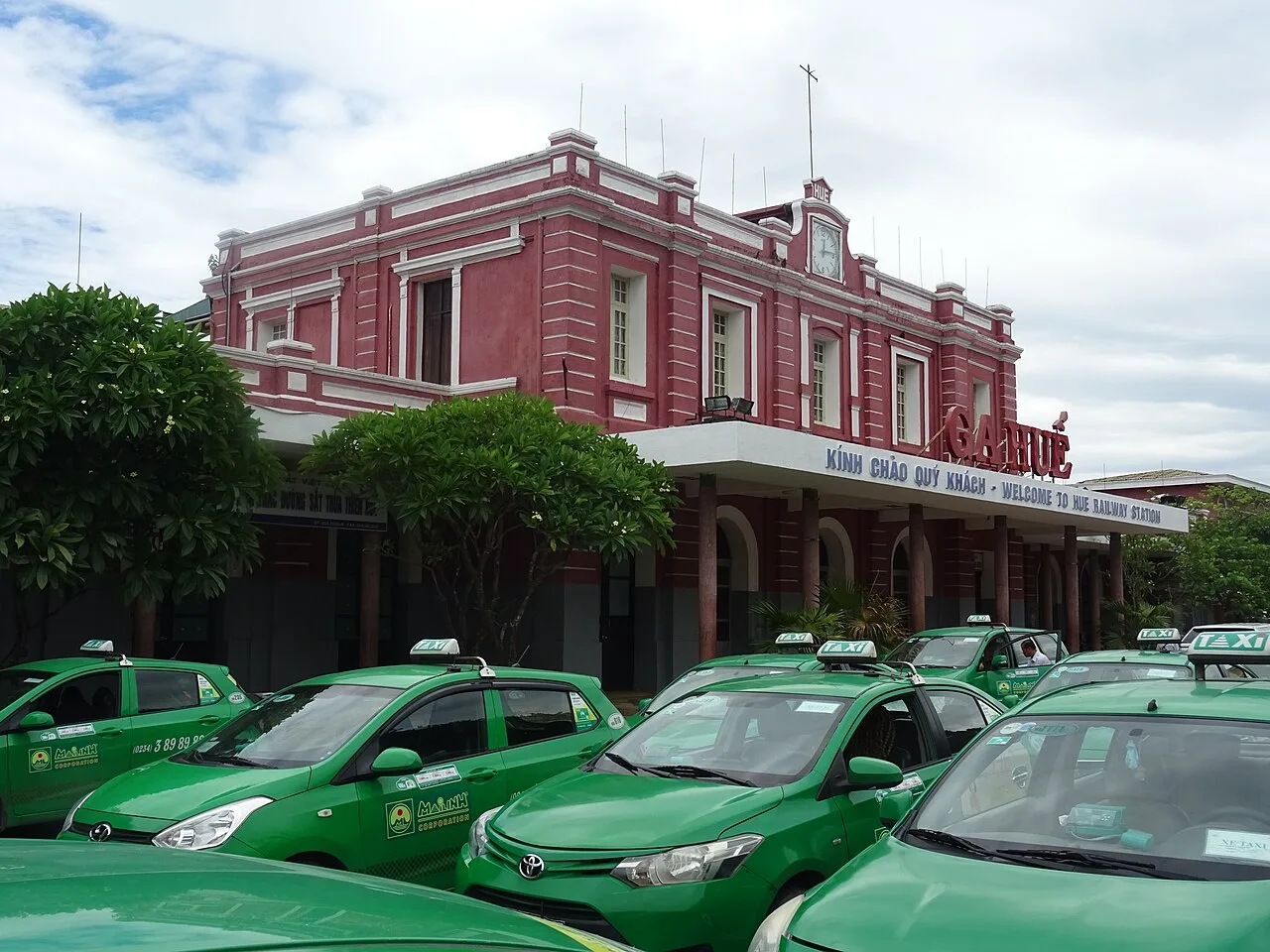 Façade rose de la gare de Huế, avec son horloge et une rangée de taxis verts devant l'entrée