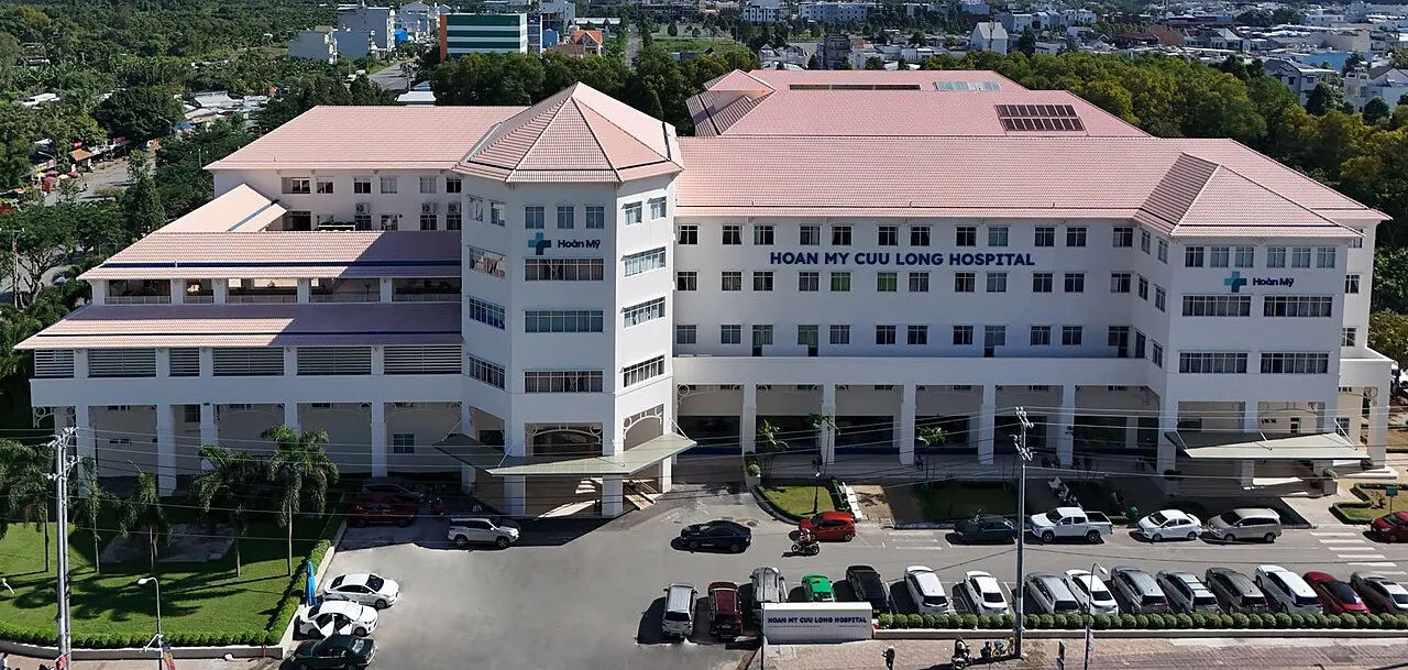 Vue aérienne de l'hôpital Hoan My Cuu Long, un grand bâtiment blanc aux toits roses, avec son parking devant l'entrée