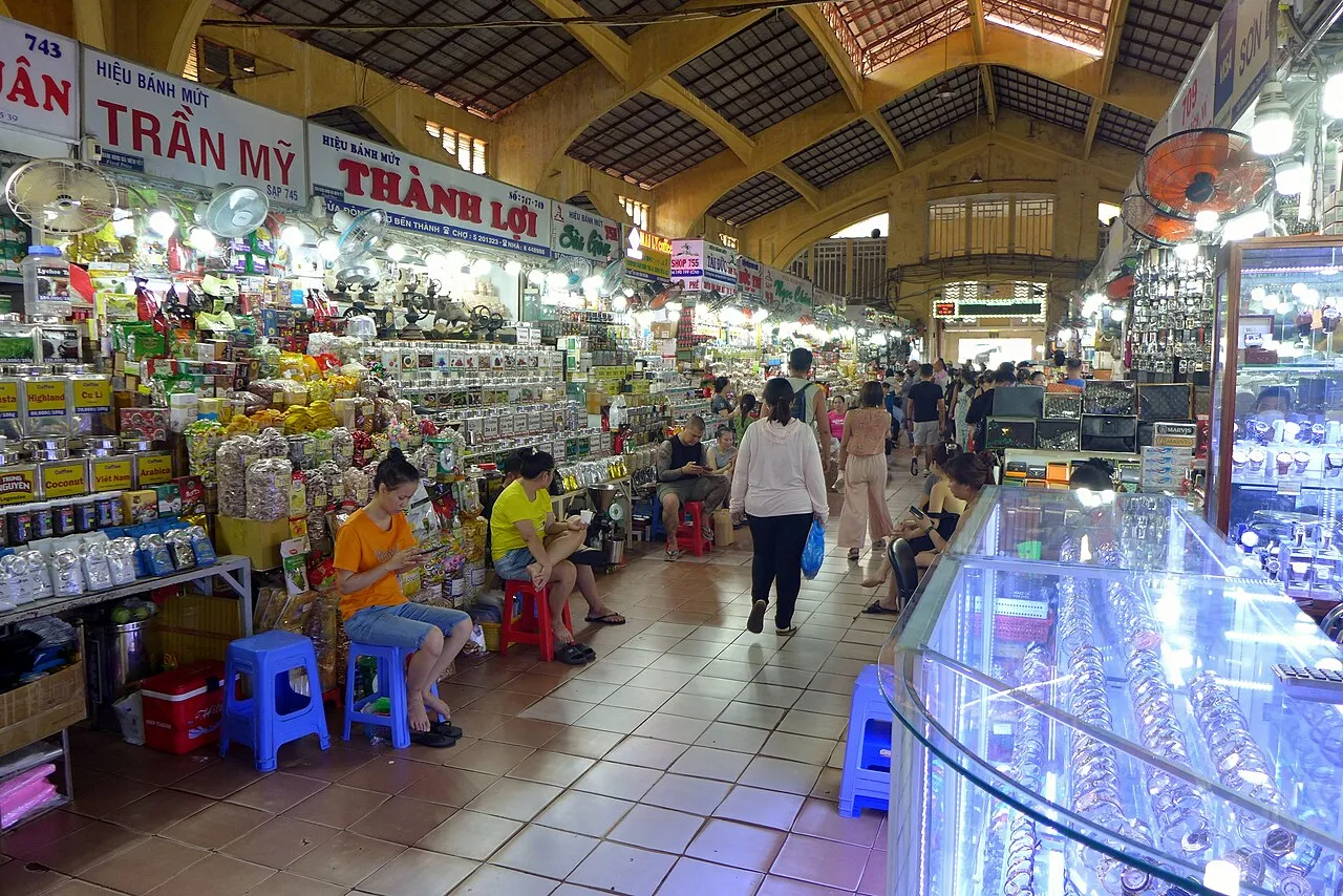 Allée intérieure du marché Bến Thành à Hô-Chi-Minh-Ville, avec des étals de produits secs à gauche, des vitrines de montres à droite et des clients qui circulent