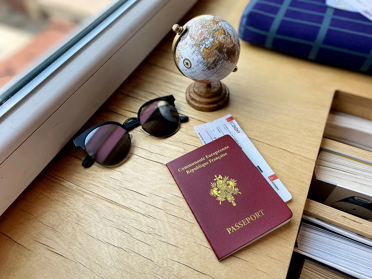 Passeport français posé sur un bureau en bois avec une carte d'embarquement, à côté de lunettes de soleil et d'un petit globe terrestre