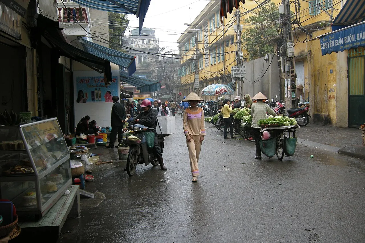 Rue commerçante du vieux quartier de Hanoï, avec des boutiques sous auvents, des passants et des vendeuses de légumes à vélo