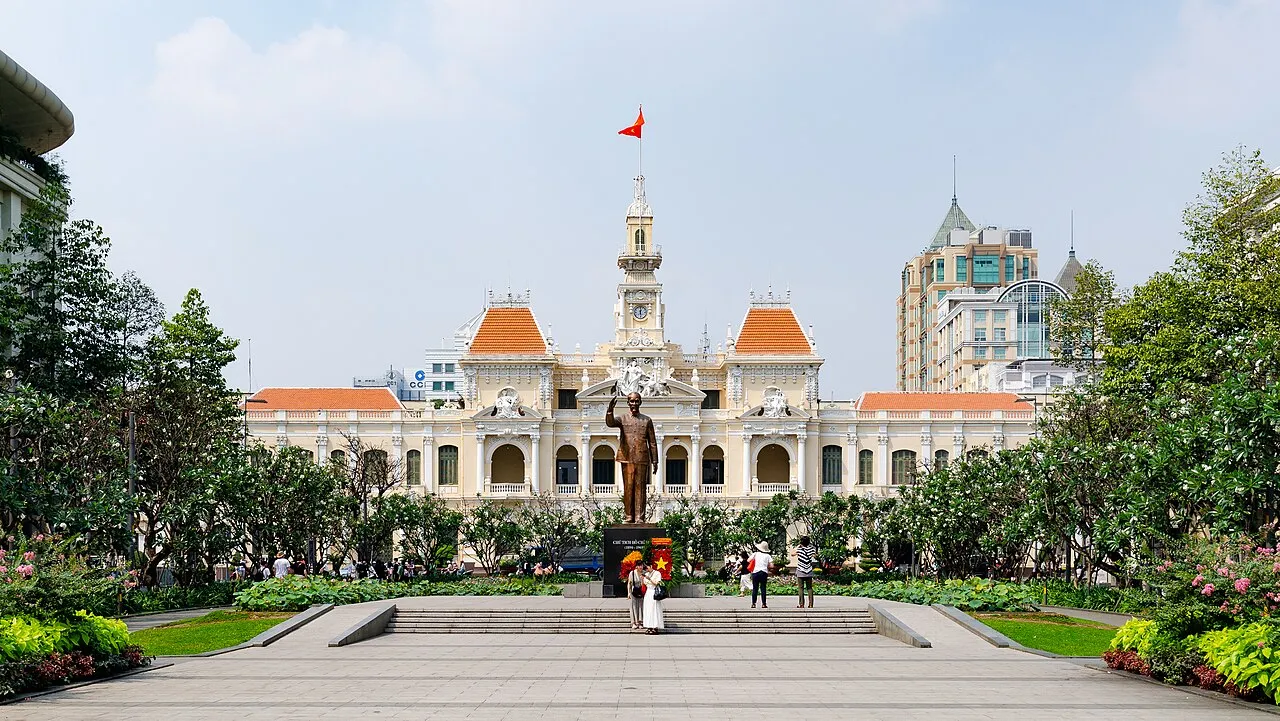 L'hôtel de ville de Hô Chi Minh-Ville, bâtiment jaune aux toits orange surmonté d'un clocher, siège du comité populaire, vu depuis l'esplanade fleurie avec une statue en bronze au premier plan