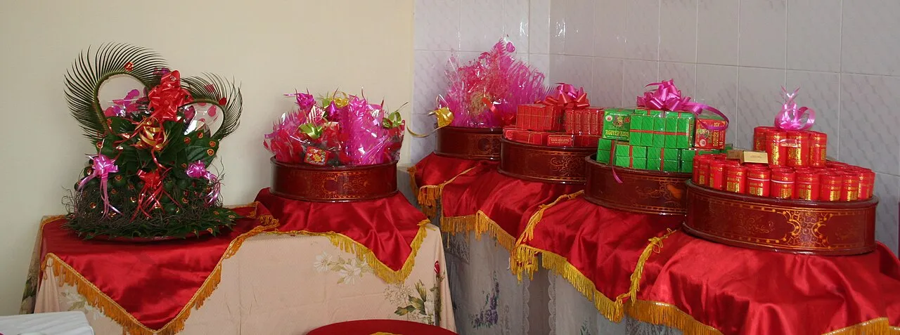Plateaux de cadeaux de fiançailles vietnamiens alignés sur des tables drapées de rouge, garnis de boîtes rouges et vertes et de cellophane rose